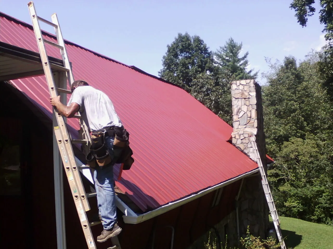 Roofer on a ladder beside a red metal roof and stone chimney — Soffit & Fascia Repair by Roofing Brothers in Tobyhanna